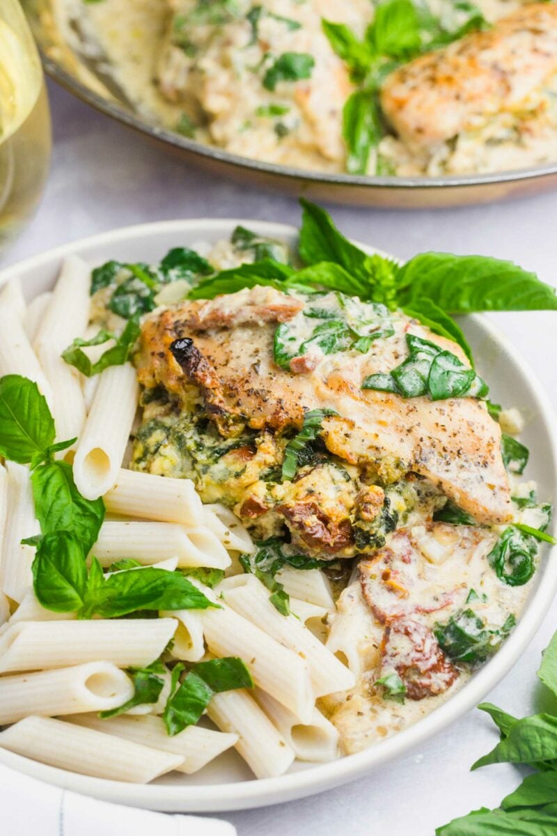Stuffed chicken breast is served in a white bowl with penne noodles and garnished with fresh basil.