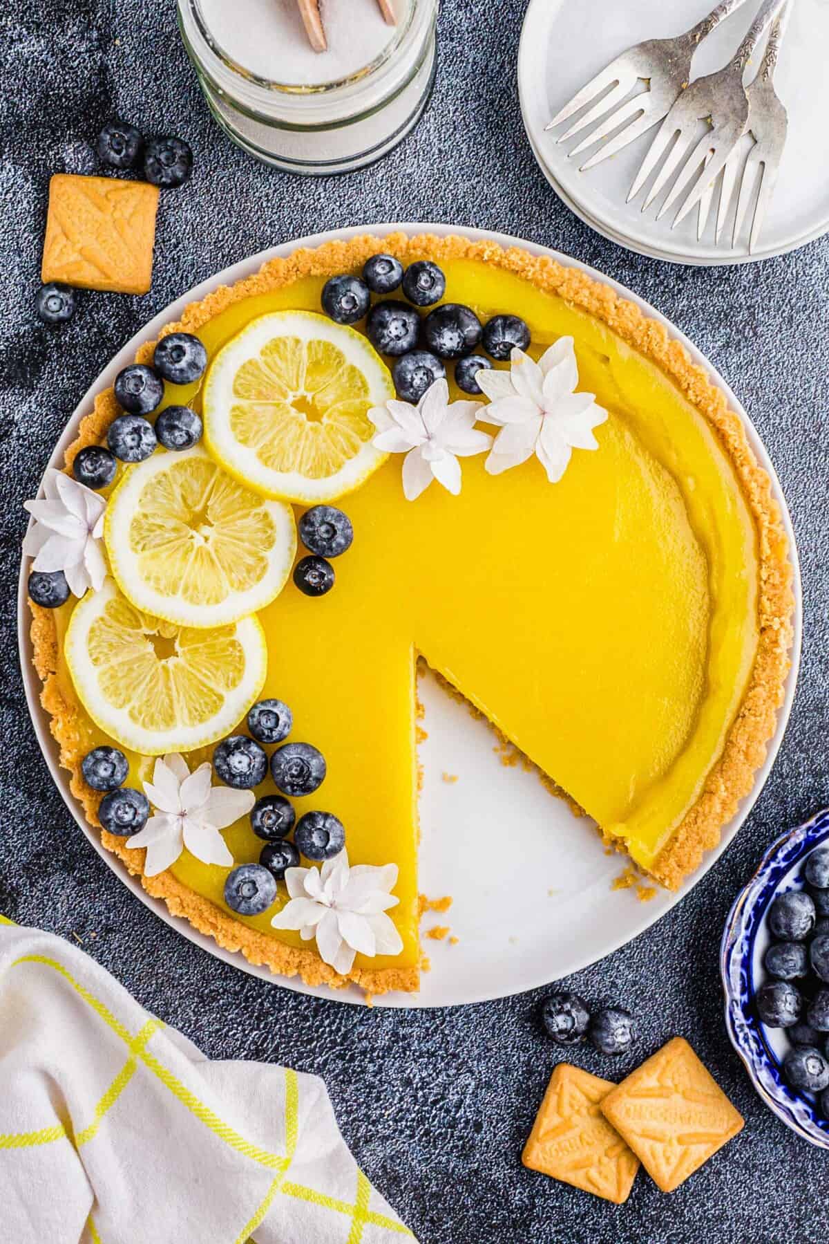 A lemon tart is garnished with lemon slices, white flowers, and blueberries.