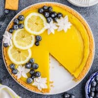 A lemon tart is garnished with lemon slices, white flowers, and blueberries.