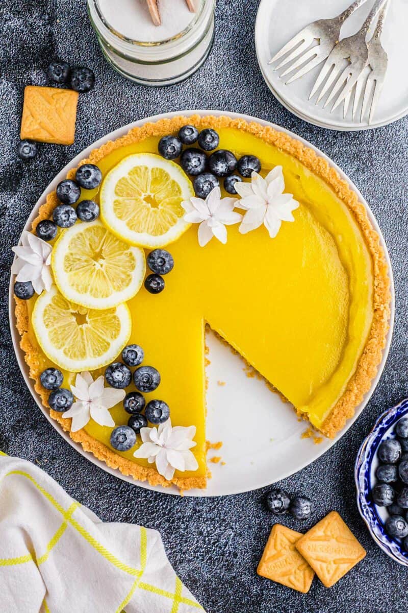 A lemon tart is garnished with lemon slices, white flowers, and blueberries. 