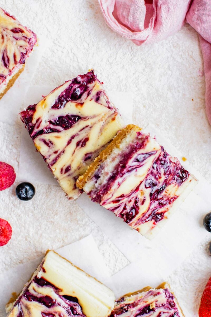 Several berry swirled cheesecake bars are placed together on a white surface. 