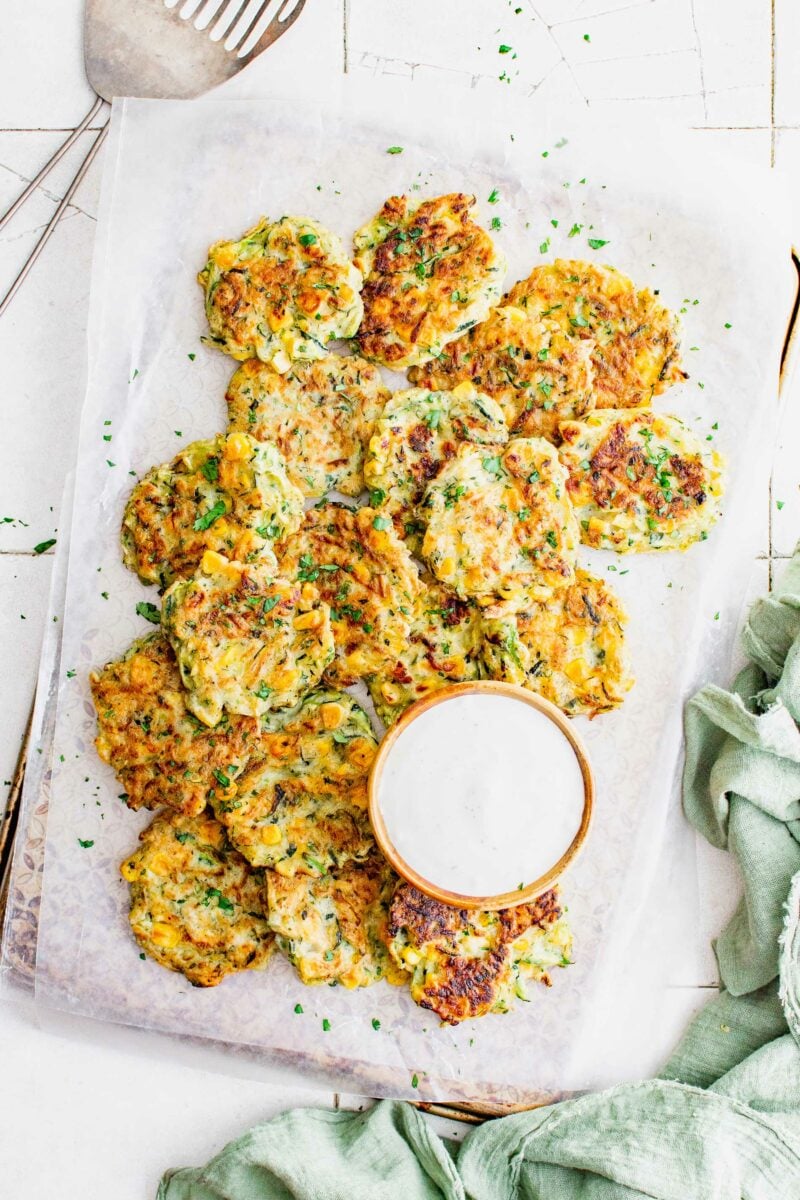 A number of fritters are placed on a prepared baking sheet next to a serving of white dipping sauce. 