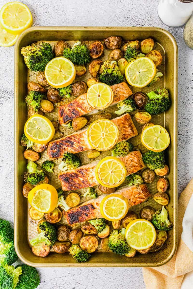 Salmon filets, lemon slices, potatoes and broccoli are spread out on a baking sheet. 