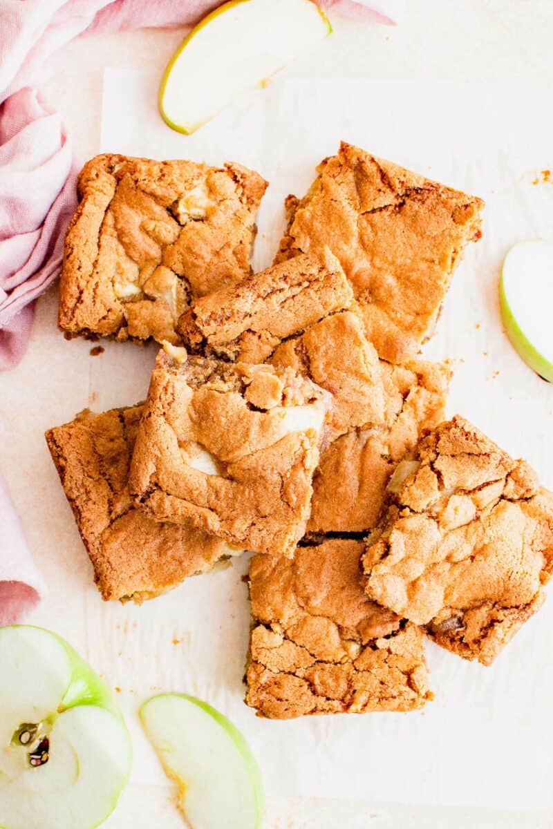 Handheld squares of apple blondies are placed on a white surface next to green apple slices. 