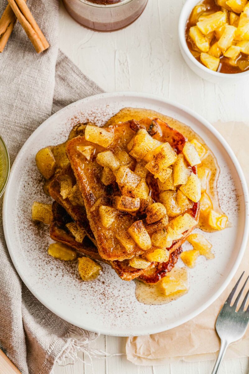 A white plate is topped with a large stack of French toast with caramelized apples on top. 