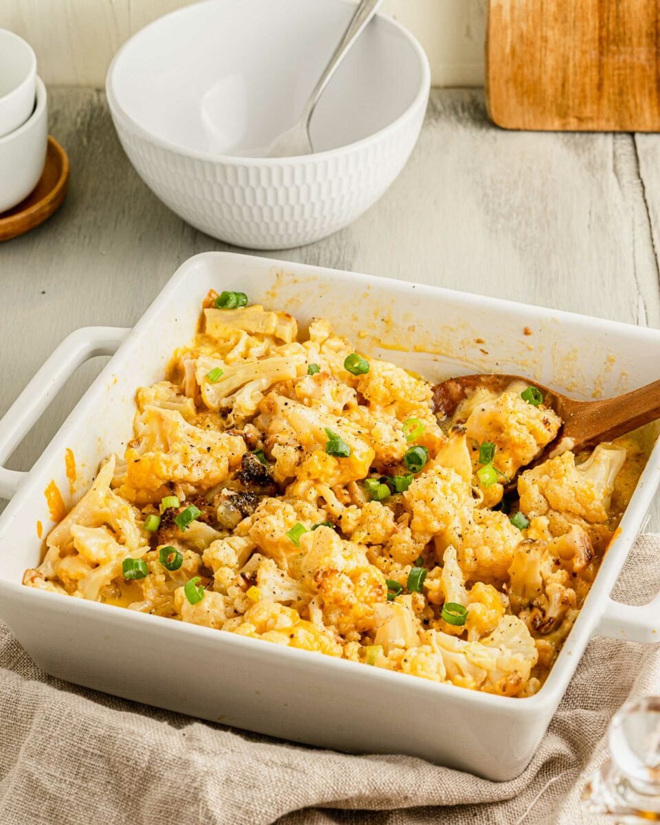 A wooden spoon is placed in a white square casserole dish filled with baked cauliflower and cheese. 