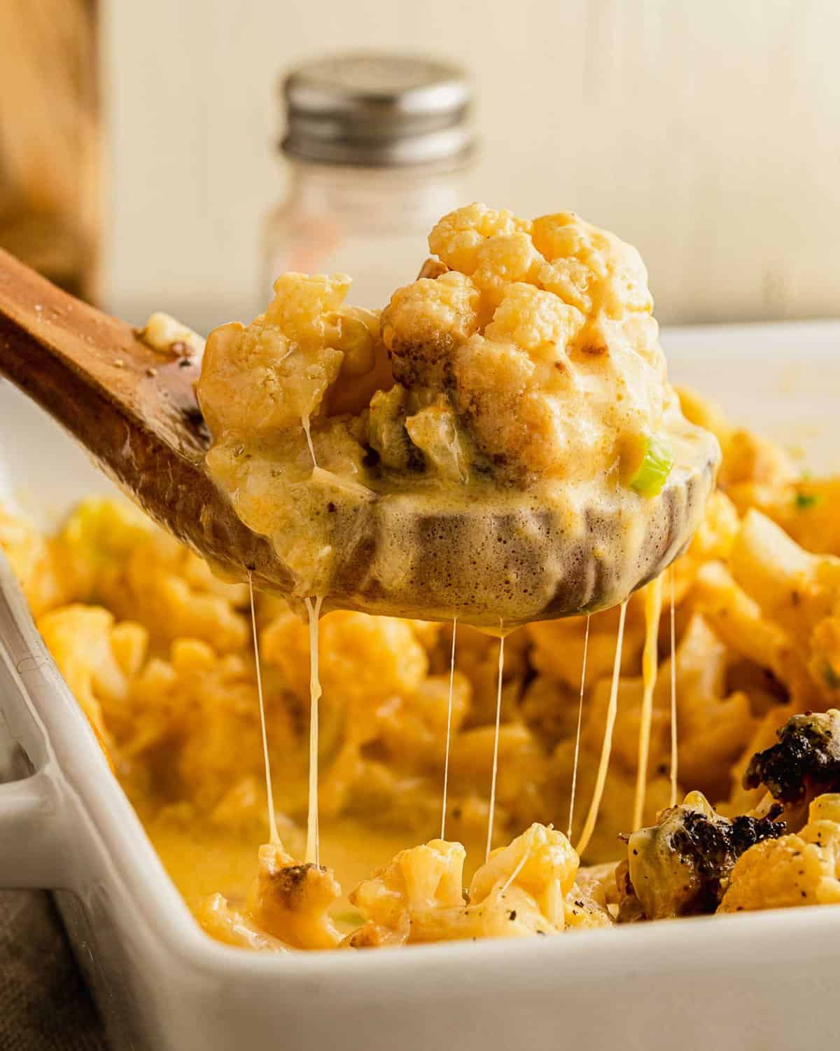 A serving of cheesy cauliflower is being lifted from the dish by a wooden spoon.