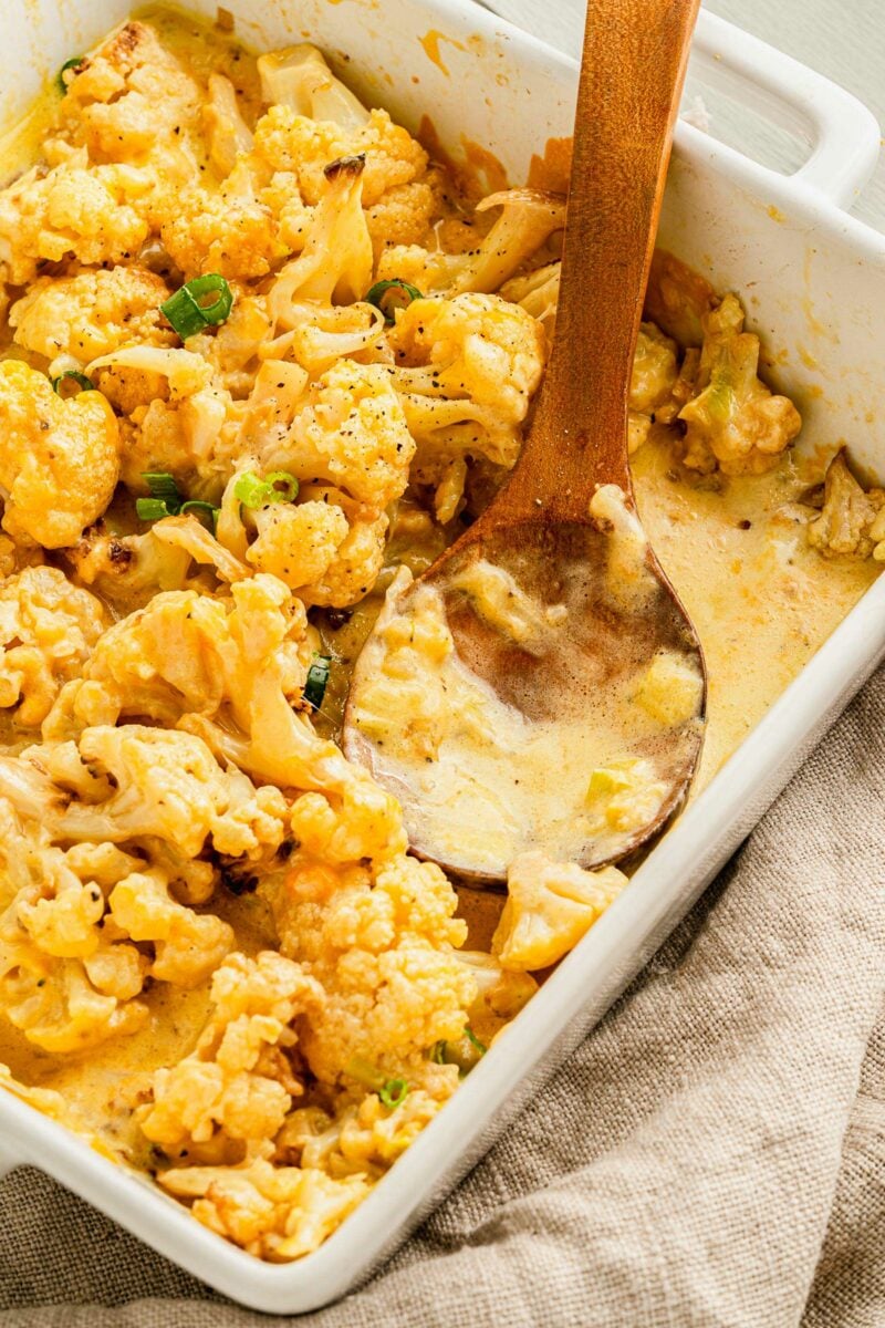 A wooden spoon is placed in a dish filled with cheesy baked cauliflower. 