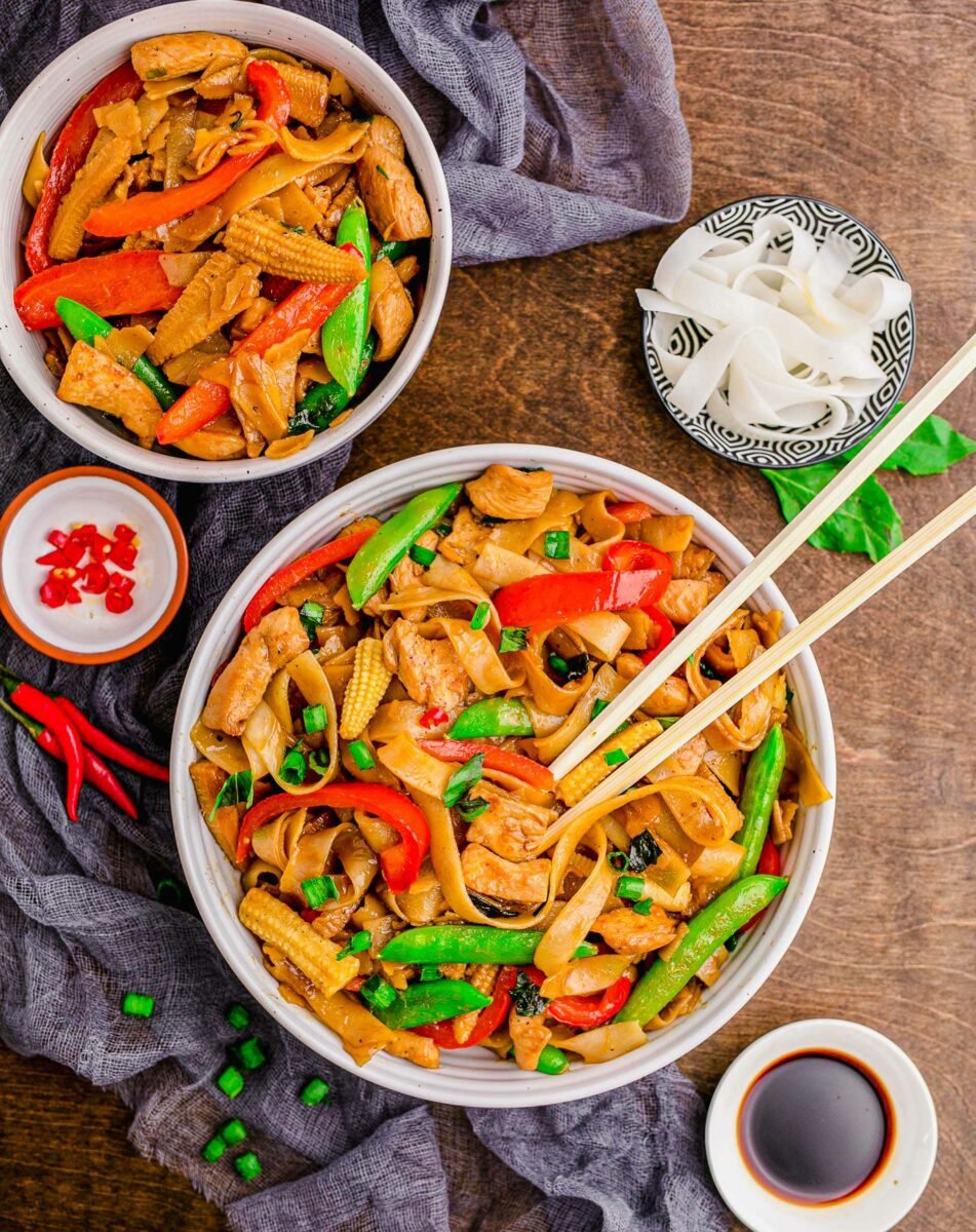 A white bowl is filled with noodles, veggies, and chopsticks. 