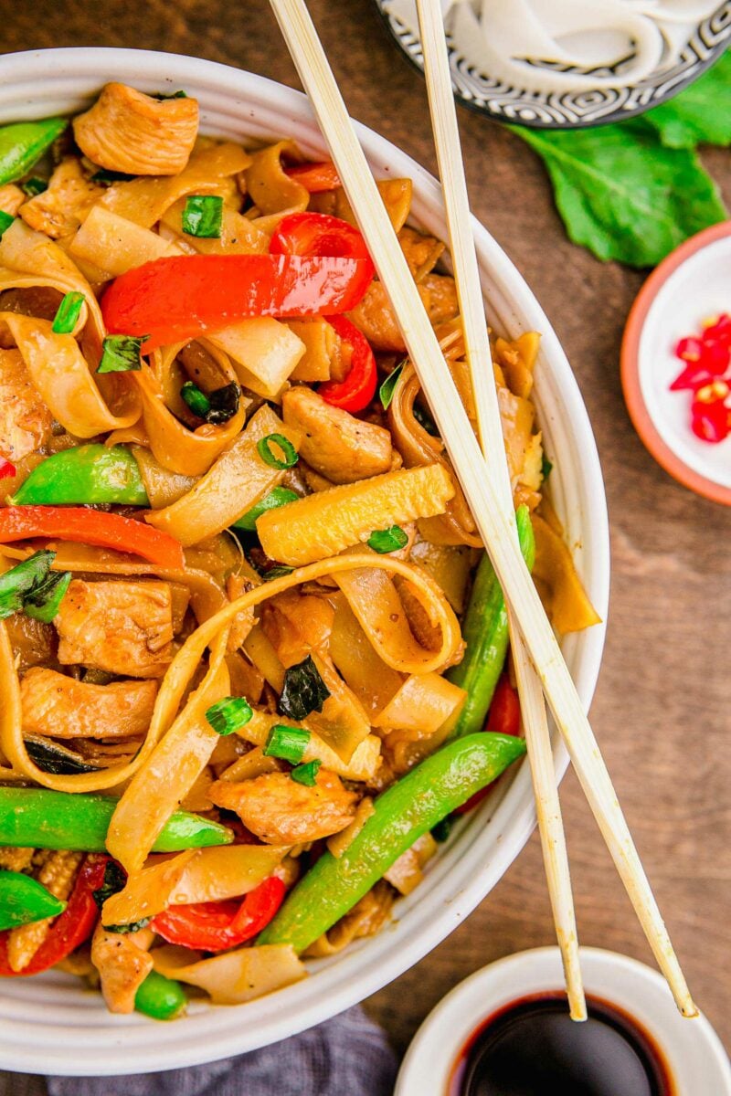 Chopsticks are placed on top of a bowl filled with noodles, chicken and veggies. 