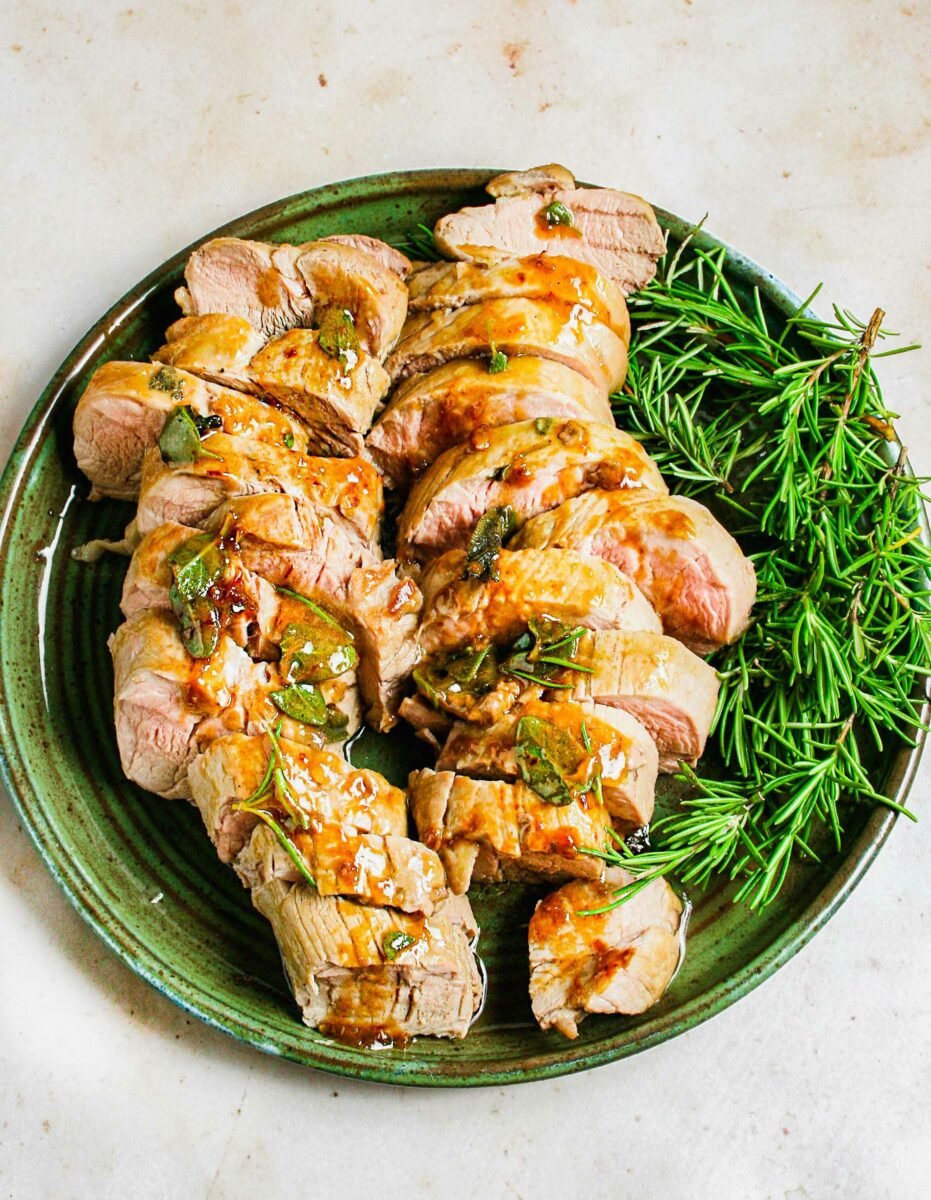 Sliced pork tenderloin is placed on a round plate next to fresh rosemary sprigs. 