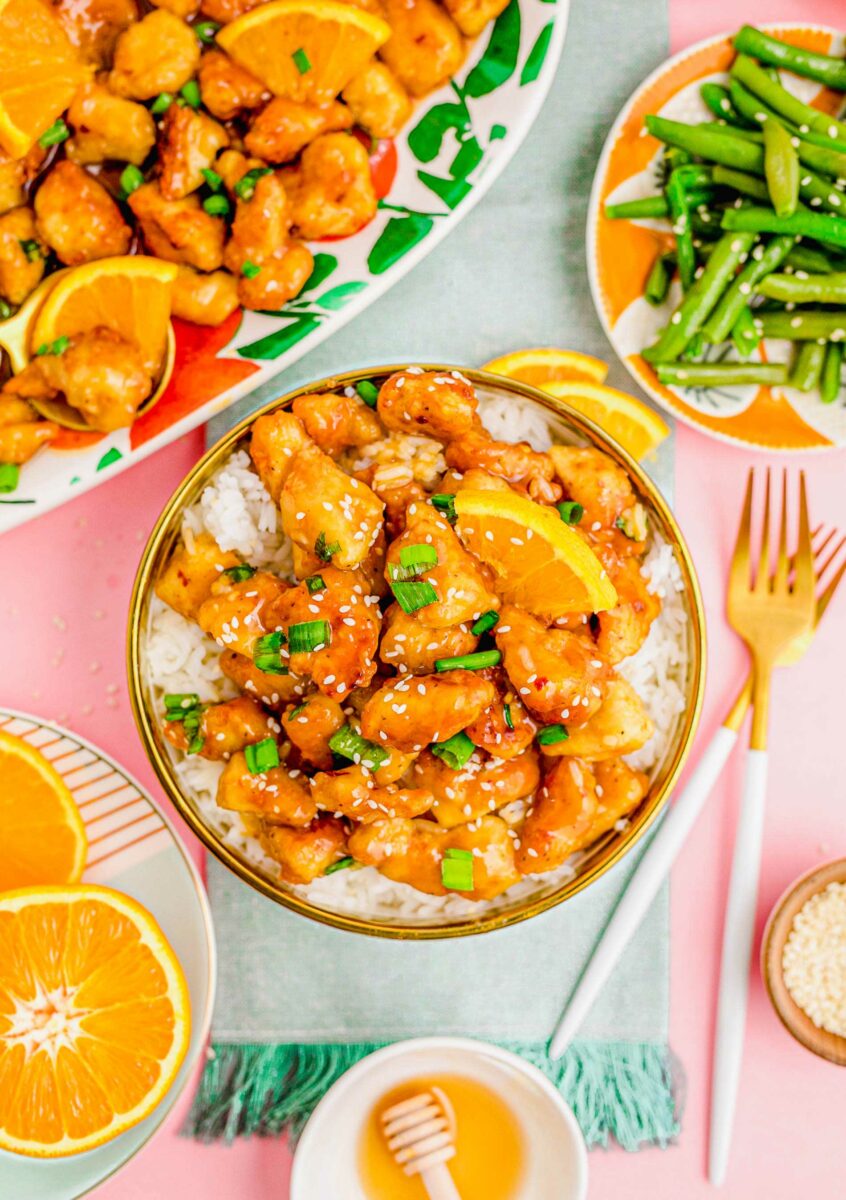 A bowl of white rice is topped with orange chicken and garnished with scallions and sesame seeds. 