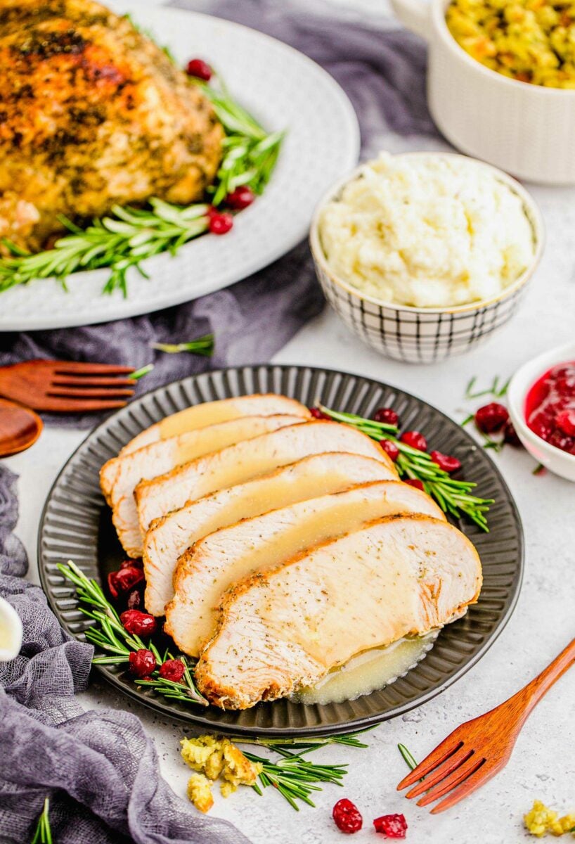 Sliced turkey breast is presented on a black plate with gravy, rosemary, and cranberries. 