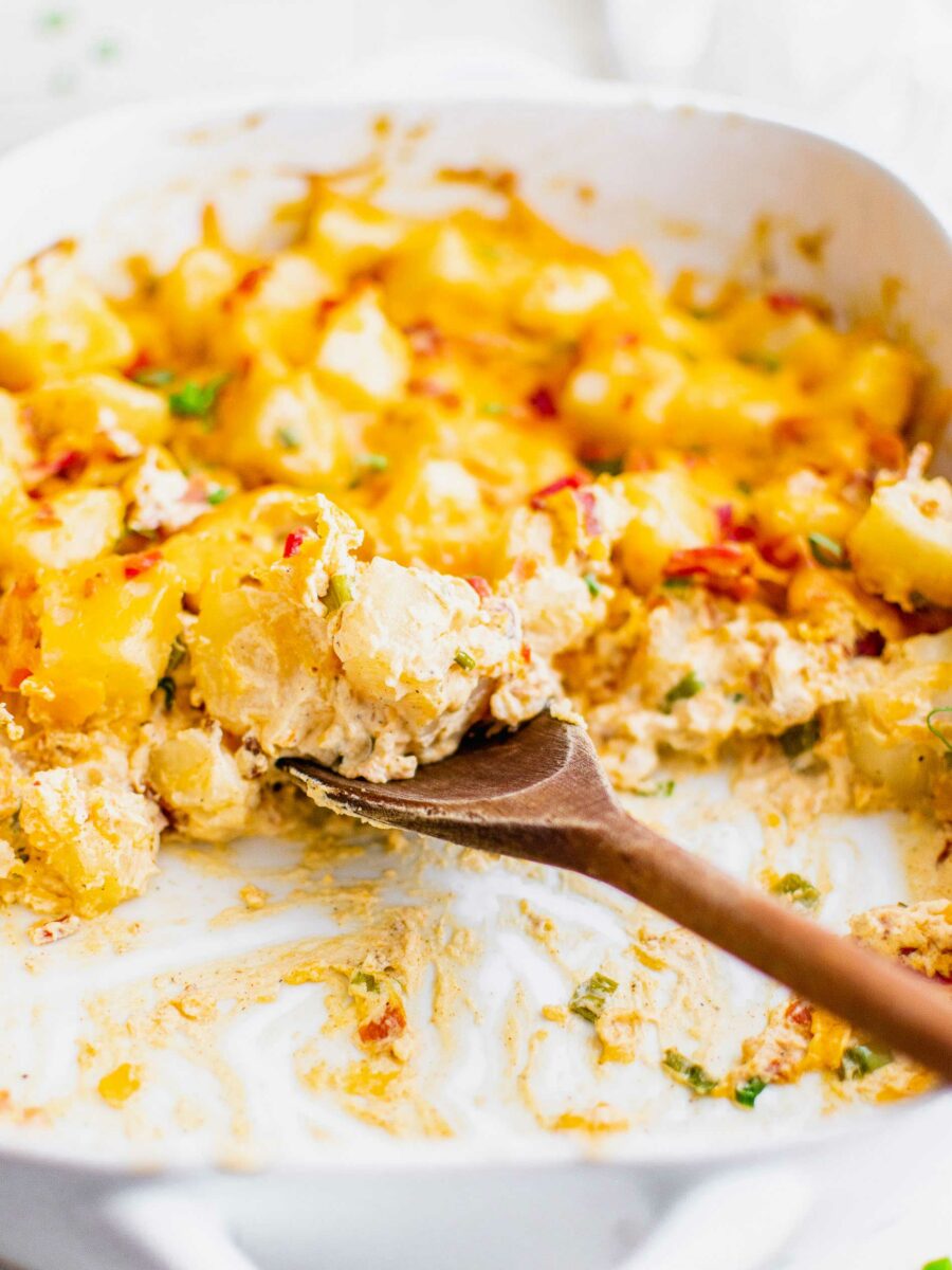 A wooden spoon is gathering a serving of potato casserole from a white dish. 