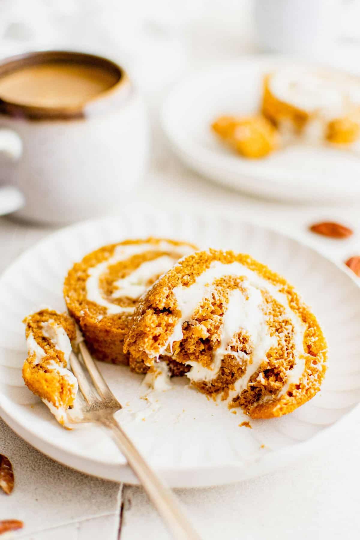 Two pumpkin roll slices on a white plate next to a fork, with a cup of coffee in the background.