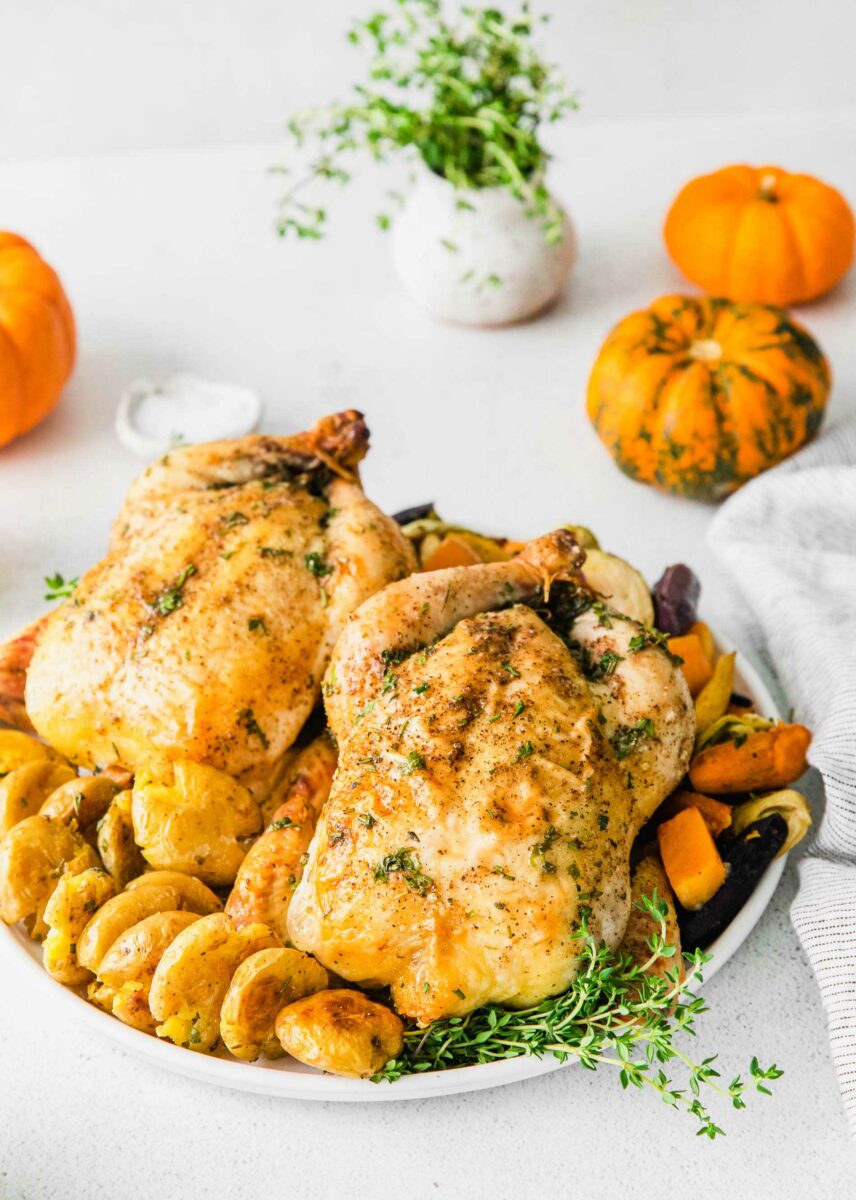 Two cornish hens are presented on a white serving plate with potatoes, root veggies and fresh herbs. 