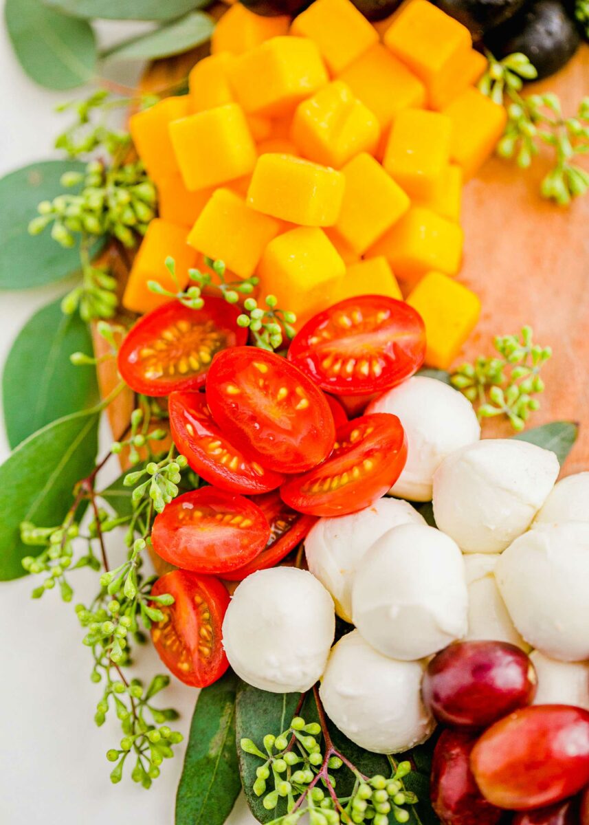 Fresh cheeses, fruits, and veggies are presented on a wooden serving platter with fresh greenery surrounding it. 