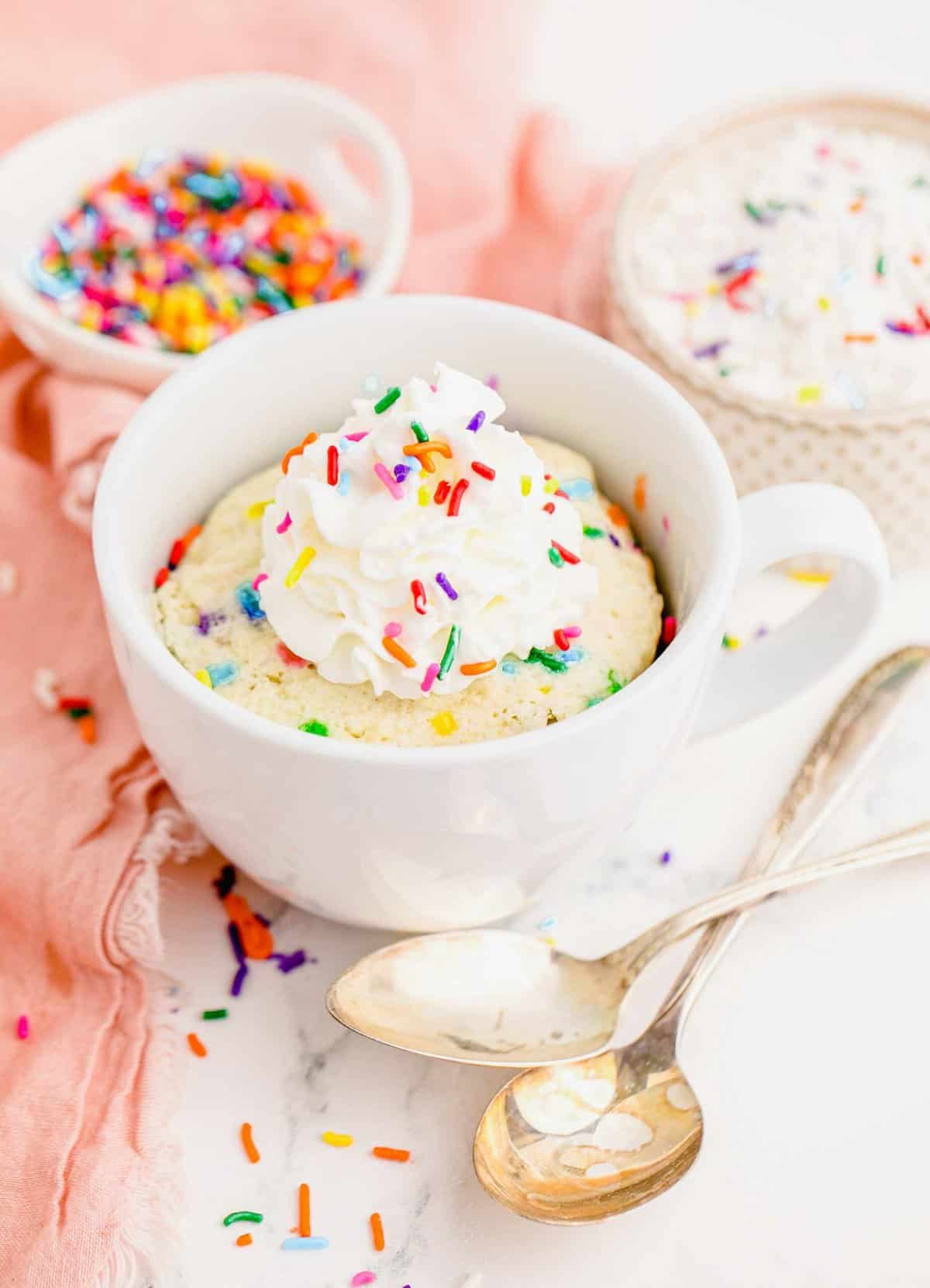 Mug of funfetti cake with dollop of whipped cream and sprinkles on top, pictured with two spoons