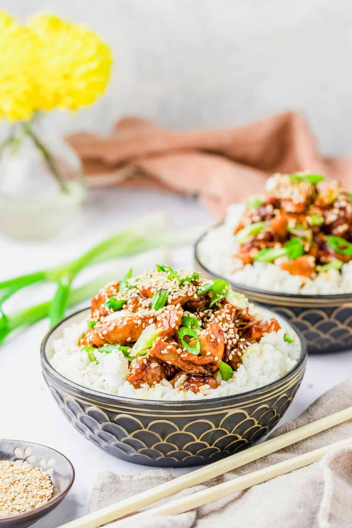 Two bowls filled with white rice and topped with honey sesame chicken, garnished with green onions