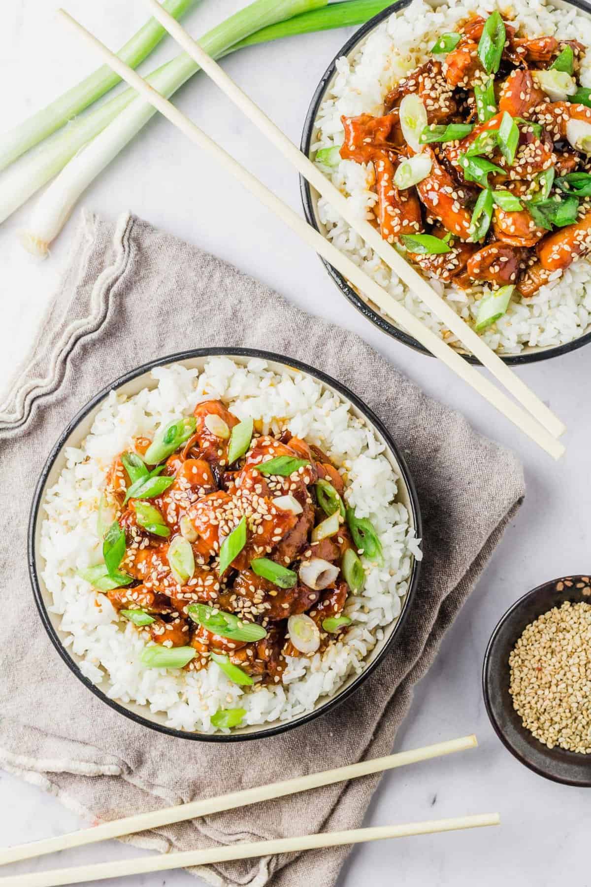 Two bowls of crockpot honey sesame chicken served over white rice, garnished with green onions