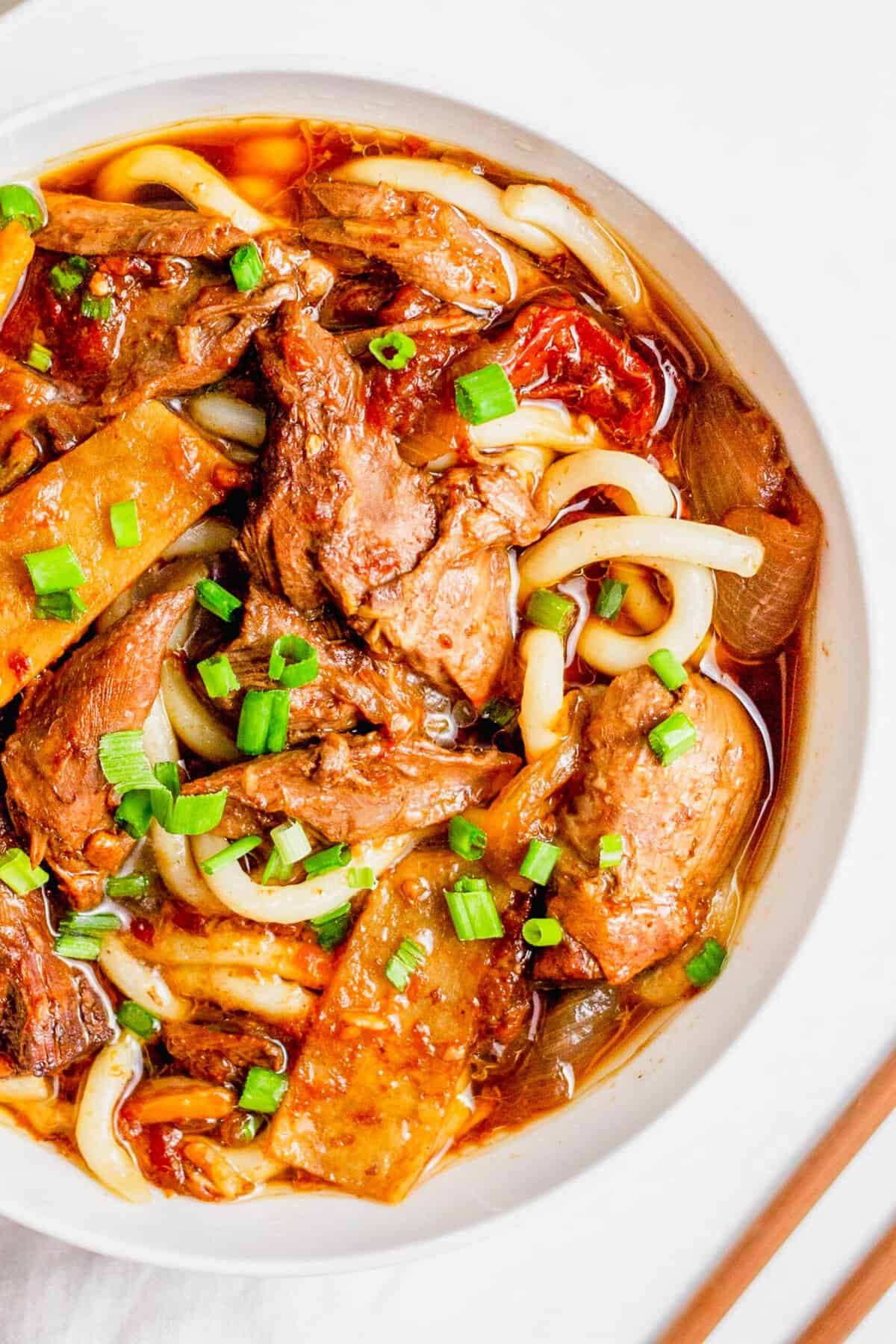 Overhead view of beef noodle soup in white bowl, topped with green onions