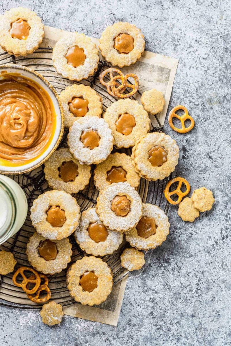 A batch of caramel pretzel linzer cookies are placed around a white bowl filled with caramel sauce.