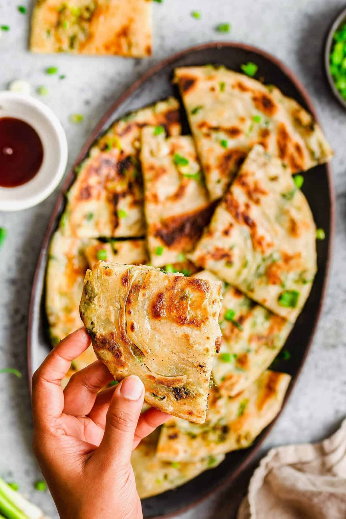 Hand holding a scallion pancake over a platter of pancakes