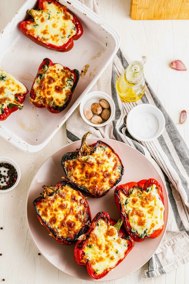 Four cooked stuffed peppers are on a round white plate next to a serving dish with more peppers. 