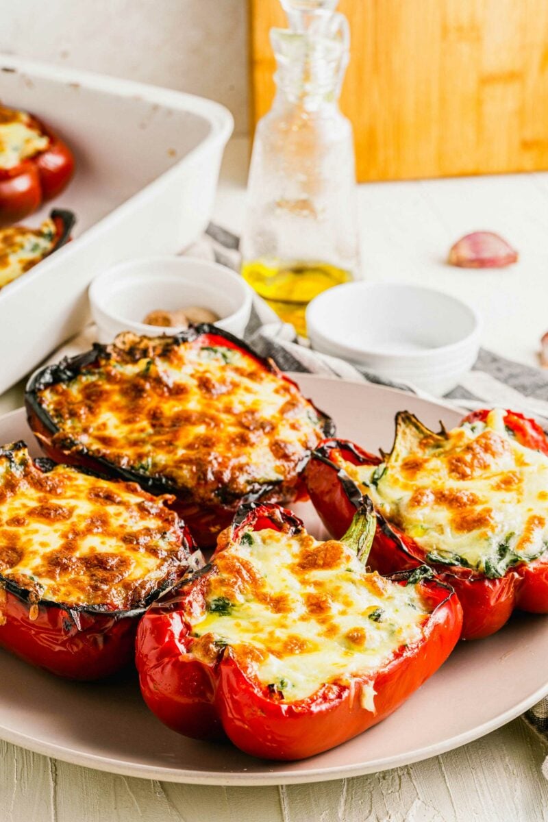 Cooked red stuffed peppers are placed on a white plate with a tea cloth in the background. 