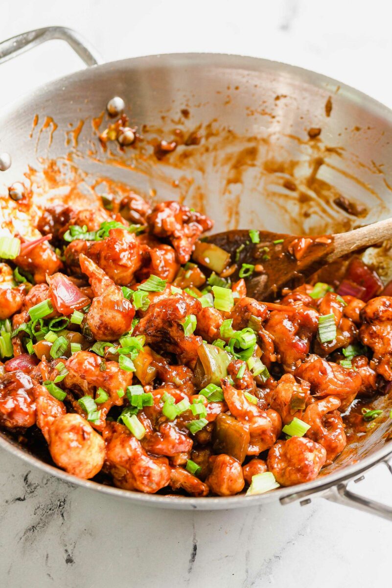 Chopped green onions are sprinkled atop a large batch of gobi manchurian in a metal pan. 