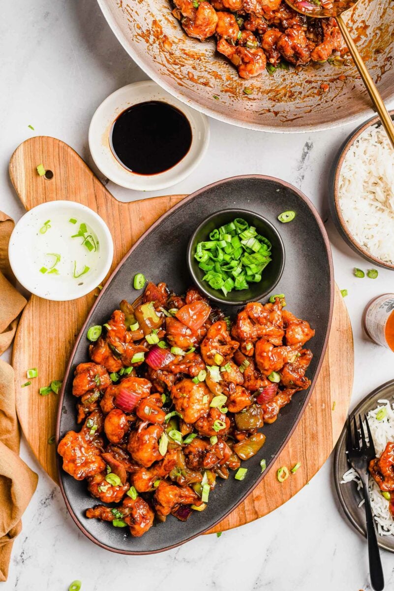 Breaded and saucy cauliflower is plated on an oval, black serving dish next to a small bowl of chopped green onion. 