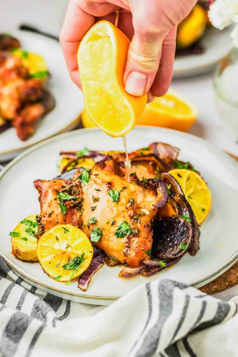 A lemon is being squeezed over a serving of cooked chicken thighs on a white plate. 