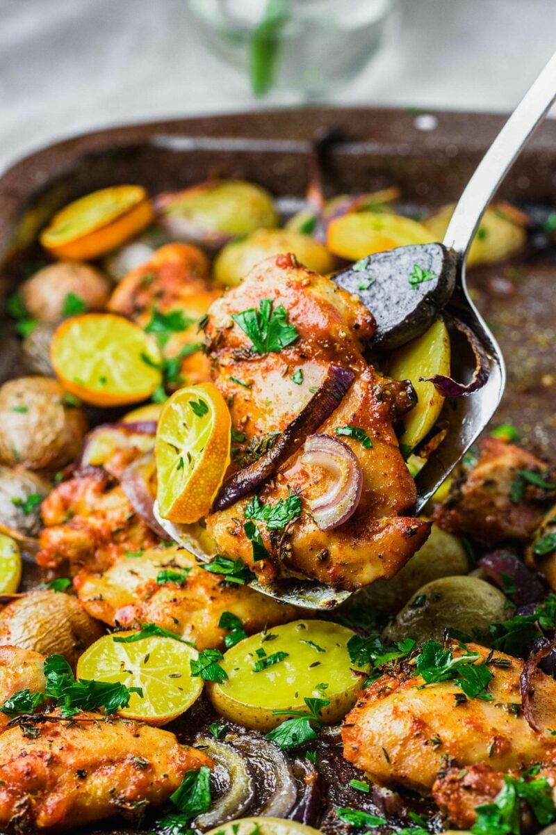 A chicken thigh with lemons, herbs, and onions is being lifted from the sheet pan. 