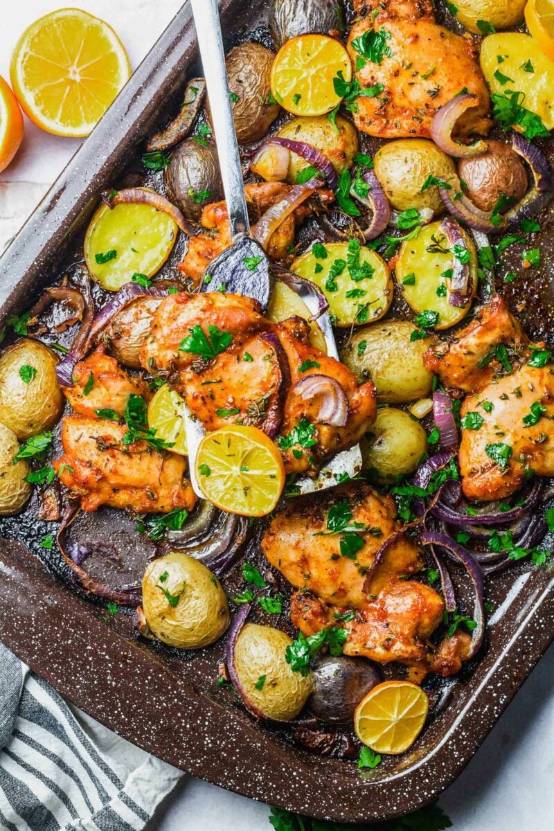 A chicken thigh is being lifted from the sheet pan with lemons and onions. 