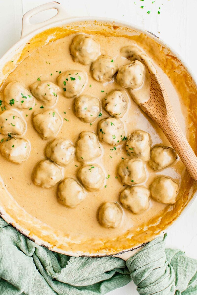 Quite a few cooked meatballs are covered in a creamy light brown sauce in a large pot with a wooden serving spoon.