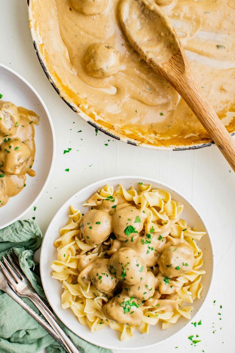 A serving of egg noodles on a white plate is topped with Swedish meatballs and fresh parsley. 