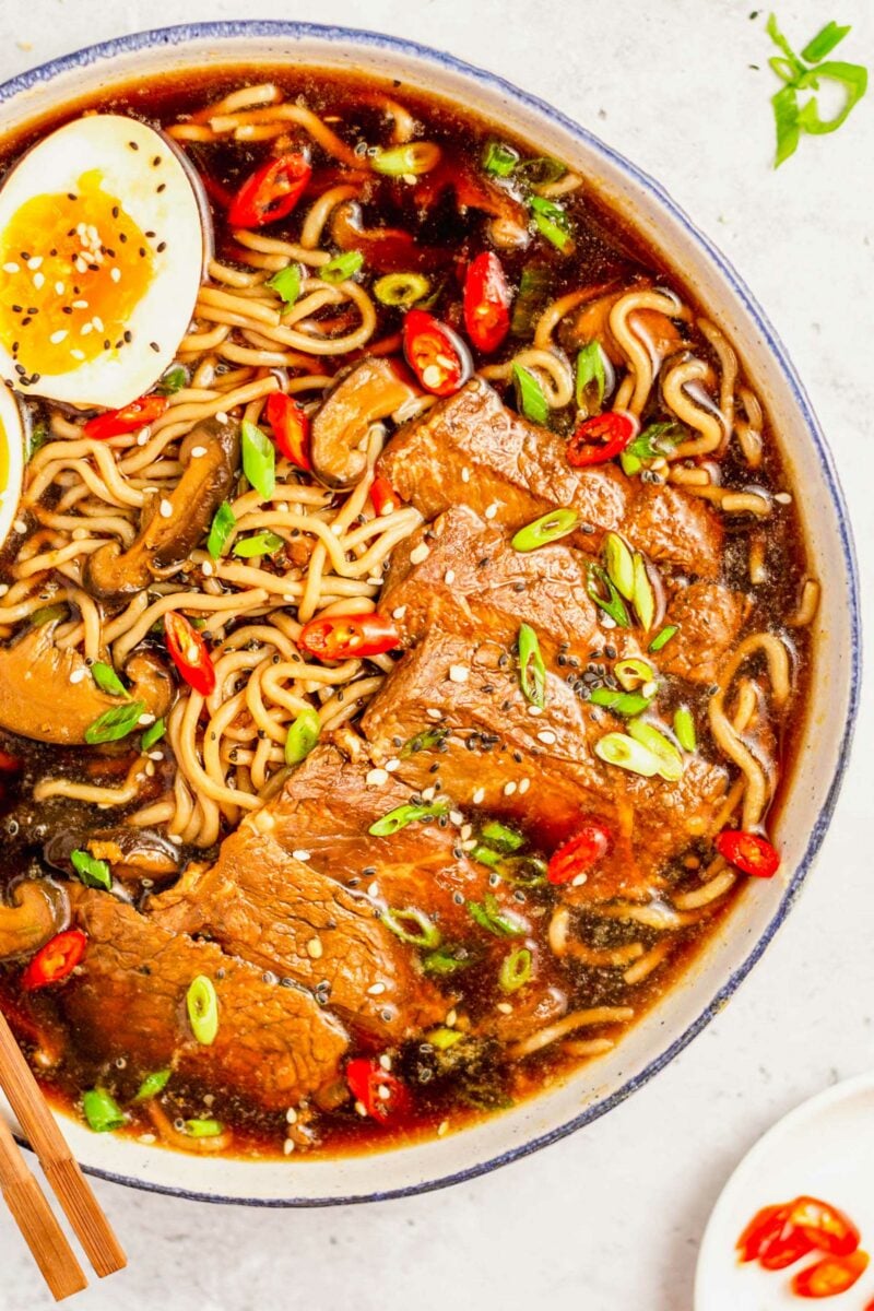 A bowl of beef ramen is garnished with freshly cut green onions and red peppers.