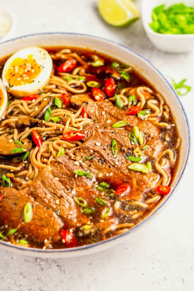 A bowl of beef ramen is garnished with freshly cut green onions and red peppers. 