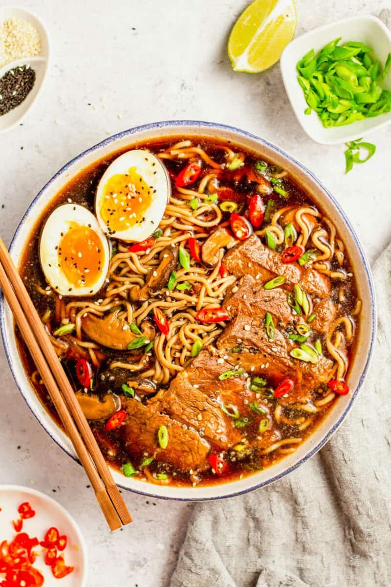 A bowl of ramen noodle soup is topped with beef, soft boiled eggs, and chopsticks. 