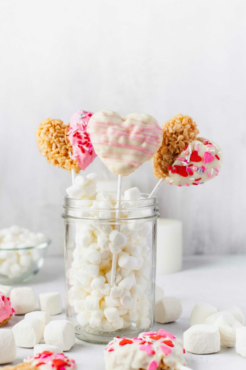 Three heart shaped rice krispie treats on white lollipop sticks are placed in a jar filled with mini marshmallows. 