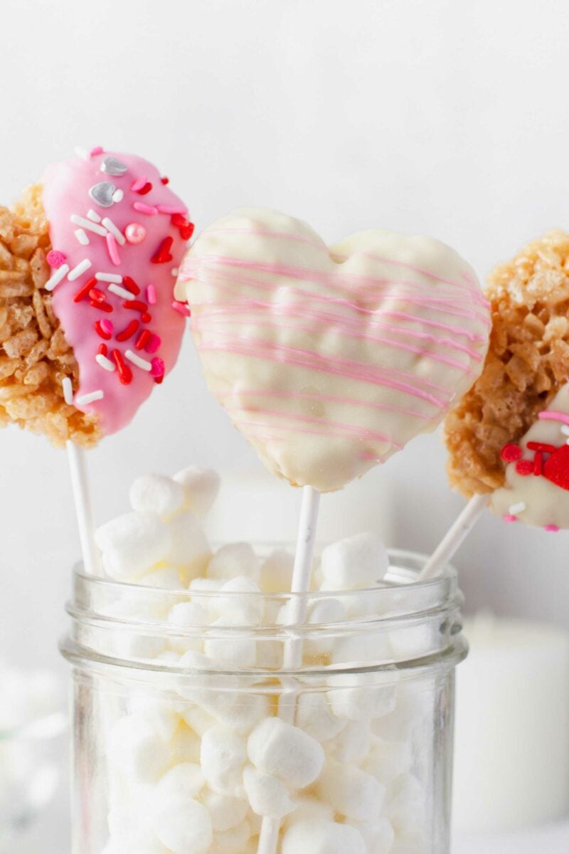 Three heart shaped Rice Krispie Treats are presented in a jar with lots of mini marshmallows. 
