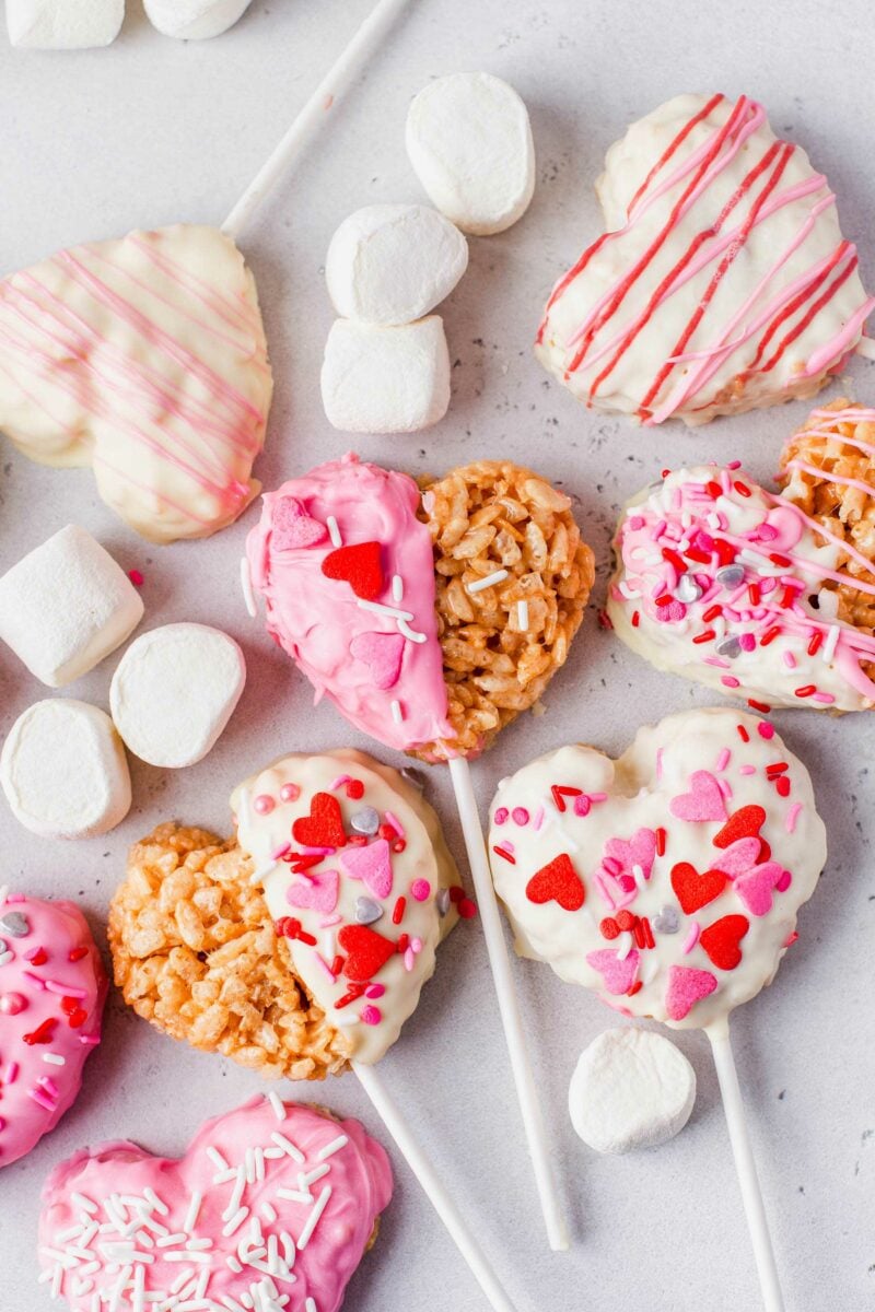 Heart shaped rice krispies treats on white sticks are placed with some mini marshmallows on a gray surface.