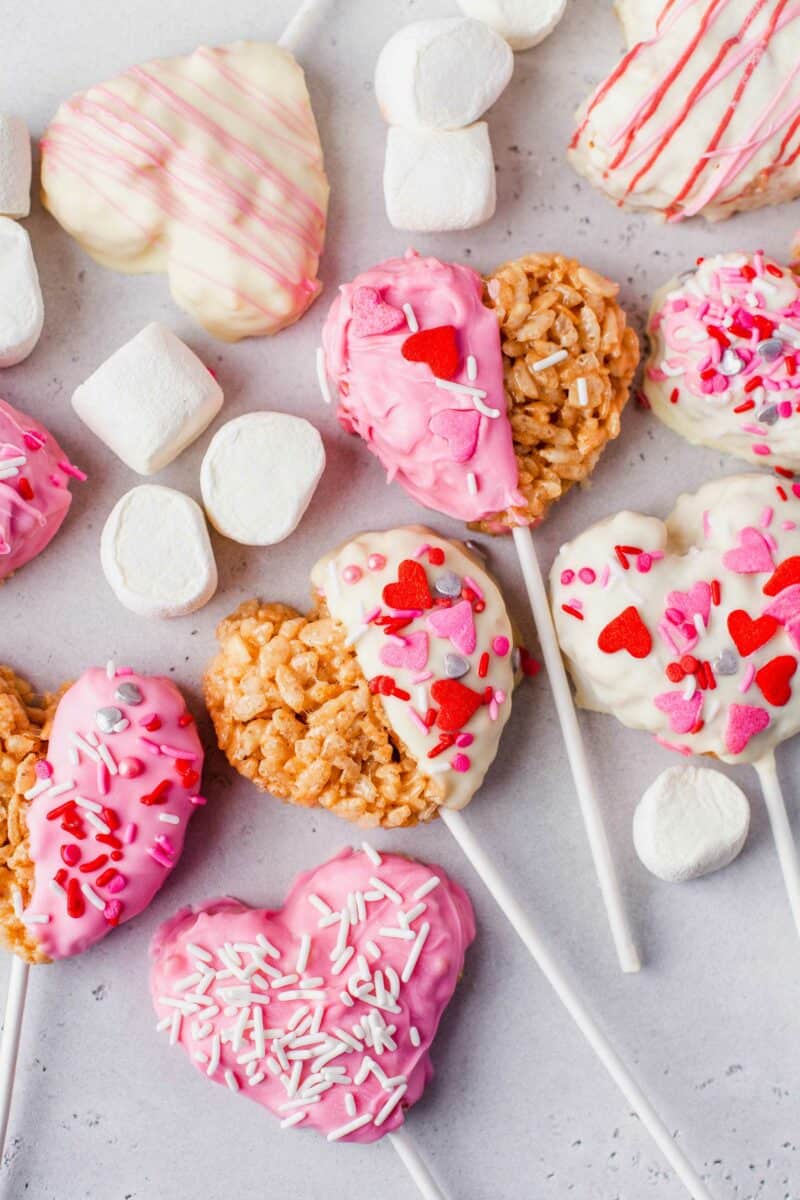 Multiple rice krispies treats hearts are presented on a gray surface with mini marshmallows. 