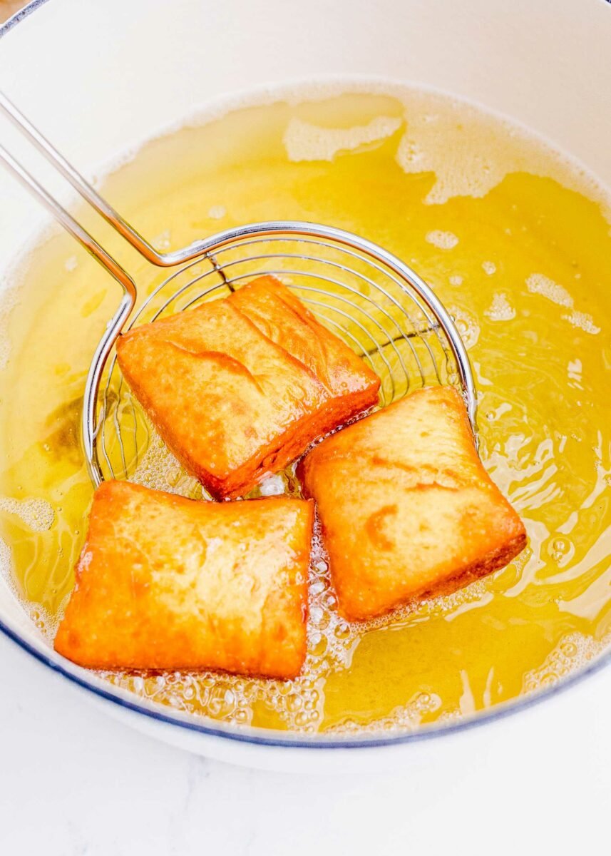 Three beignets are being fried in a large pot full of yellow vegetable oil. 
