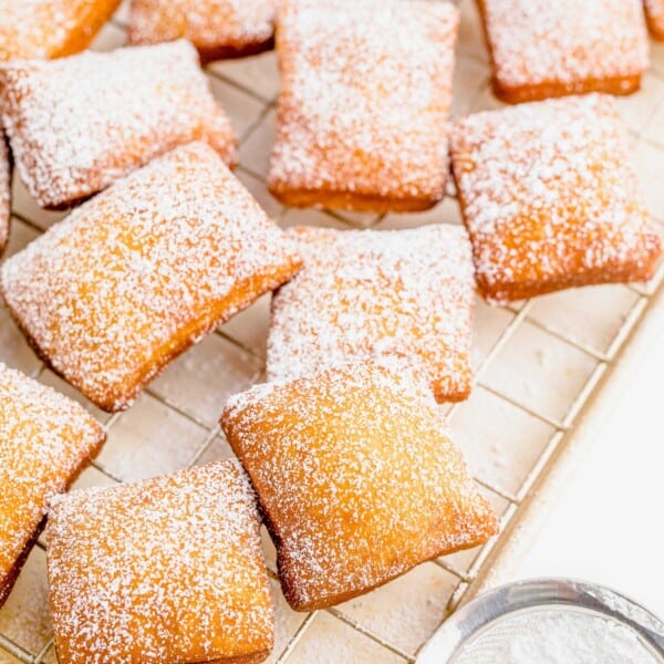 A large batch of beignets are placed on a wire cooling rack and have been dusted with powdered sugar.