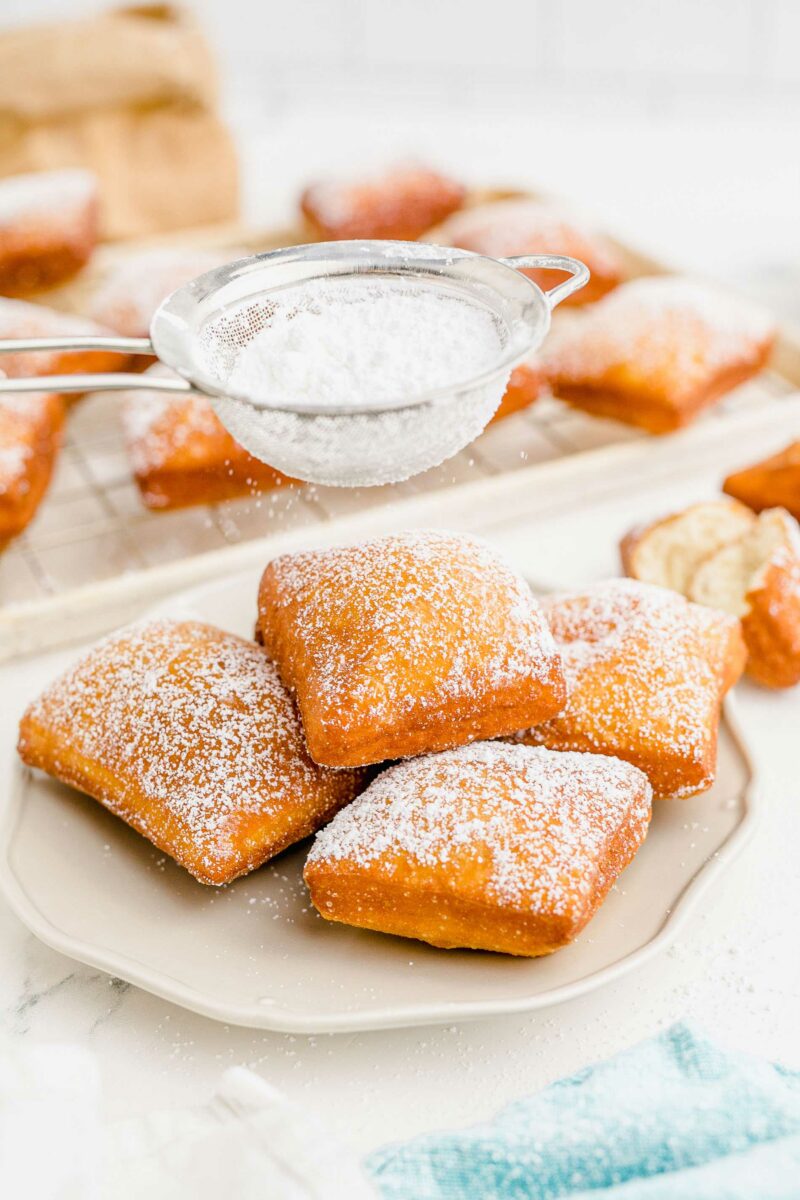 Powdered sugar is being sprinkled over a plate of beignets. 