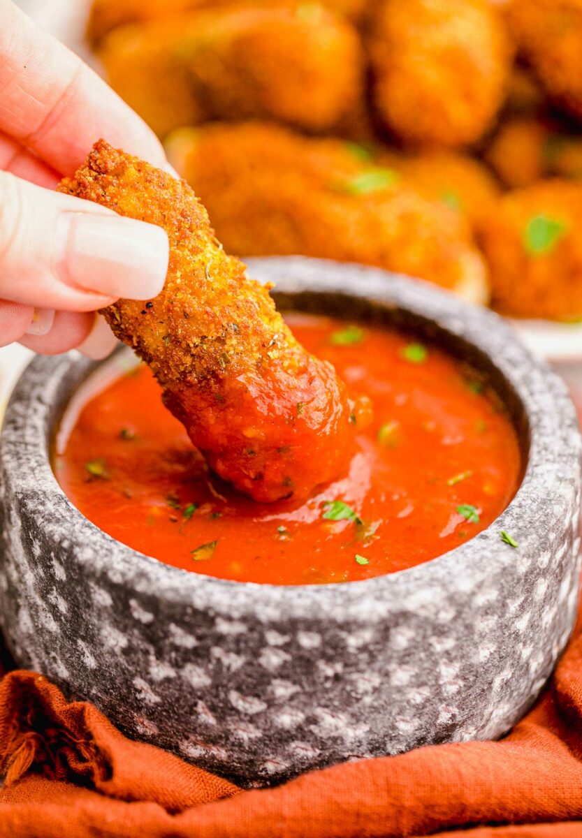 A mozzarella stick is being dunked into a bowl of red marinara sauce. 