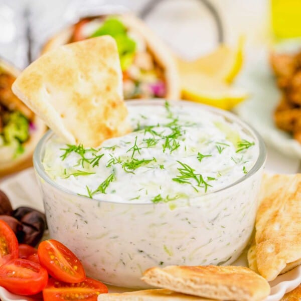 Tzatziki dip in glass bowl on plate with pita chips, olives, and halved grape tomatoes