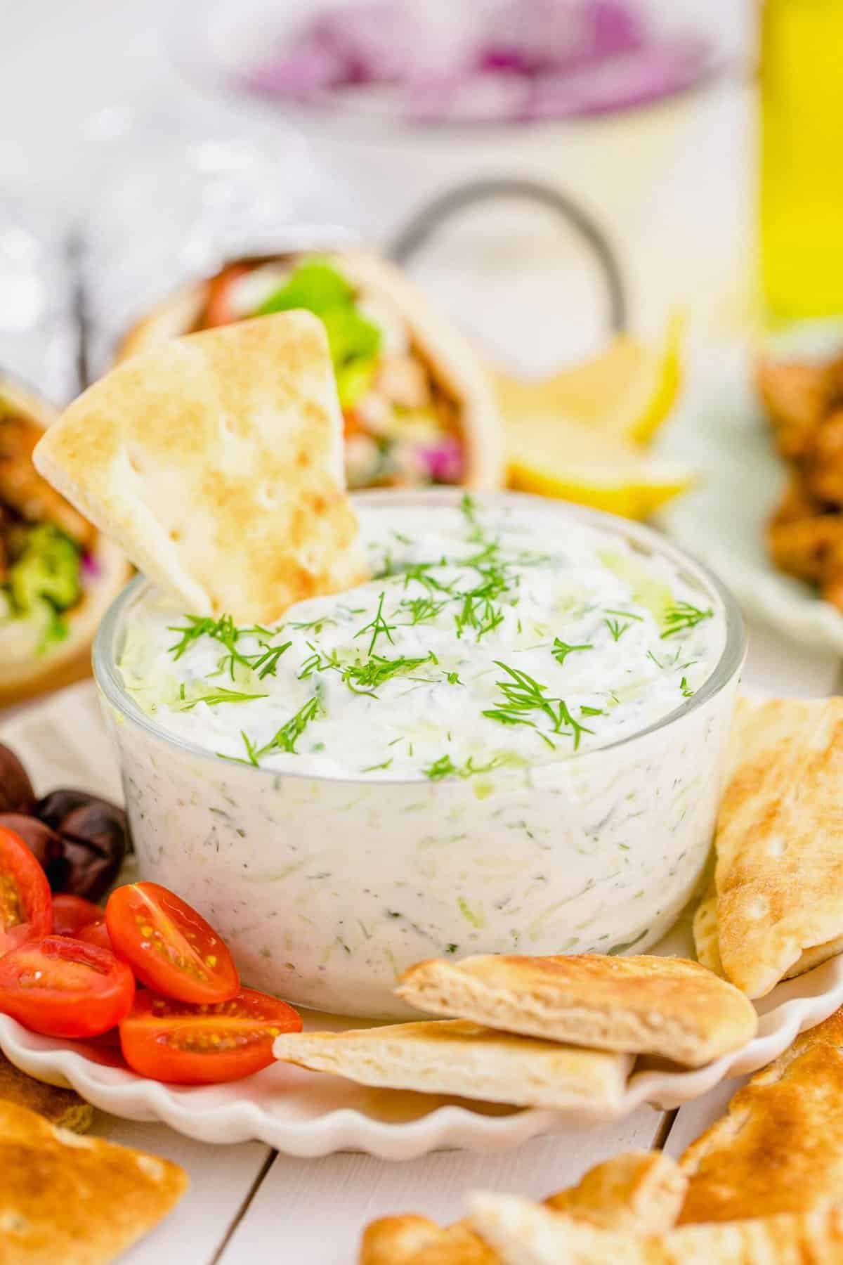 Tzatziki dip in glass bowl on plate with pita chips, olives, and halved grape tomatoes