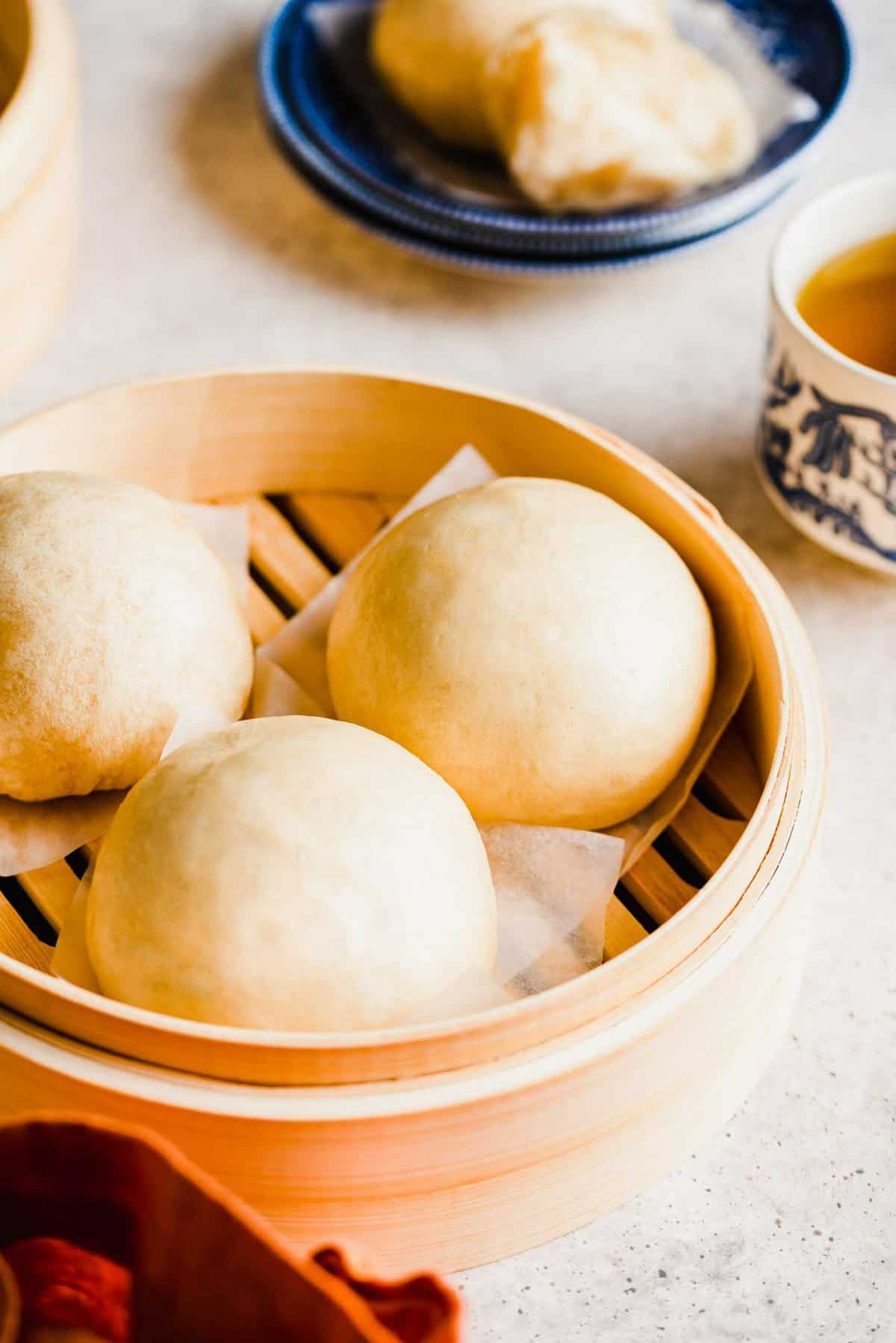Three Chinese steamed buns sitting in a steamer, with a bun on a plate in the background, next to a cup of tea